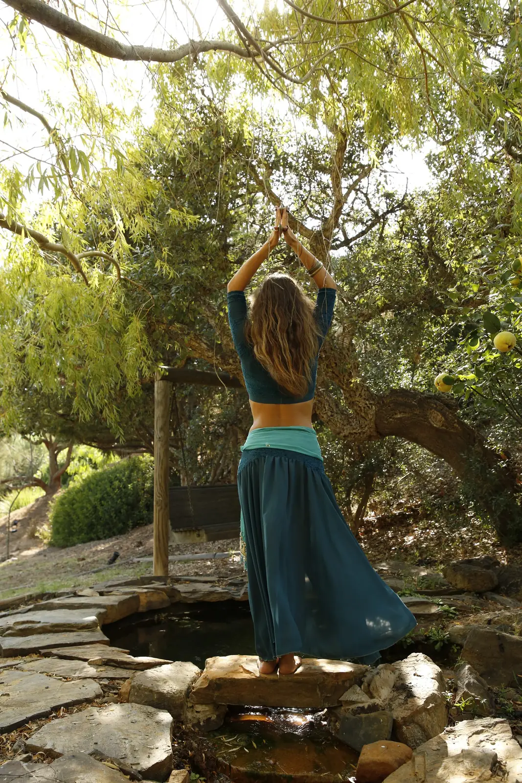 Ana de costas, de pé numa pedra junto a um pequeno lago, com as mãos unidas acima da cabeça, sob as árvores