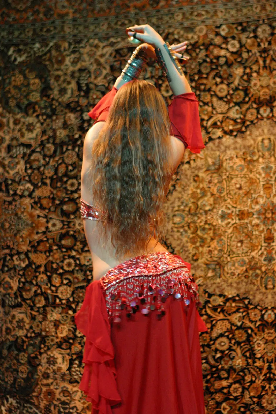 Ana de costas, com cabelo comprido ondulado e traje vermelho de dança do ventre, de braços erguidos diante de um tapete oriental